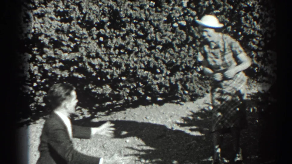 The suited man crouches or leans toward a woman in a plaid dress and white hat during the playful garden exchange.