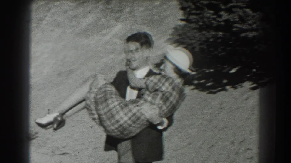 In 1939 San Francisco home-movie footage, a man carries a woman in a plaid skirt and white hat along a sunlit dirt path.
