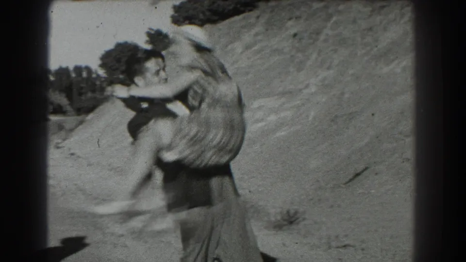The man appears to spin while carrying the woman, whose plaid skirt and white hat stand out against the bright hillside.