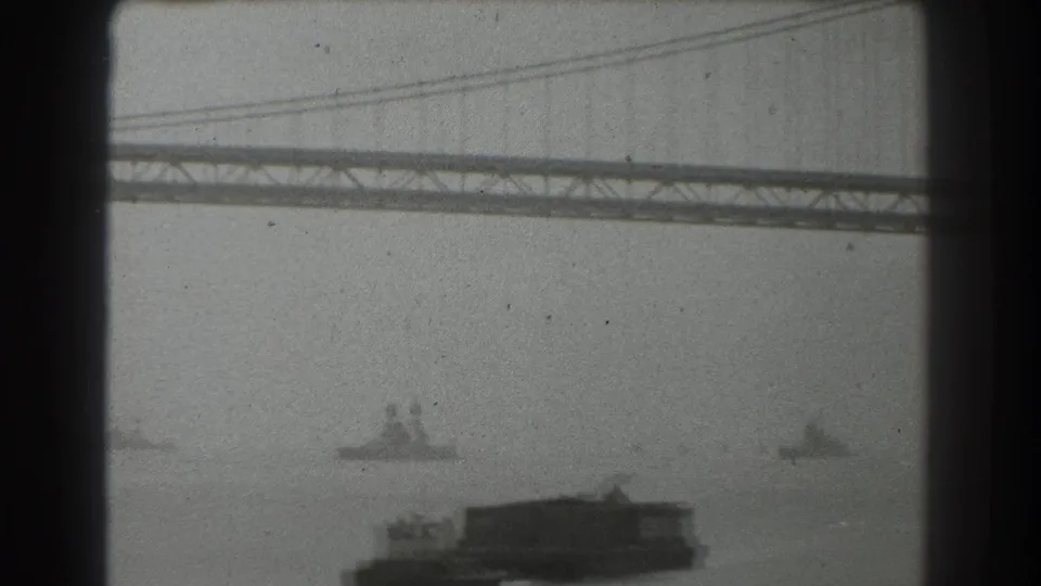 San Francisco Bay Bridge overhead with a large barge-like vessel in the foreground and smaller ships in the misty distance.