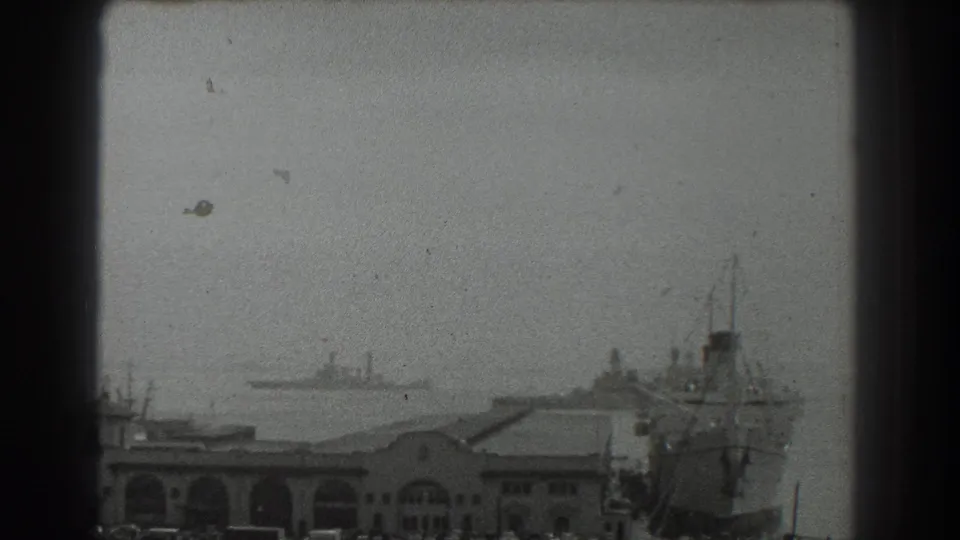 1939 San Francisco harbor footage frame with arched waterfront structures, distant ships, and a large vessel at the pier.