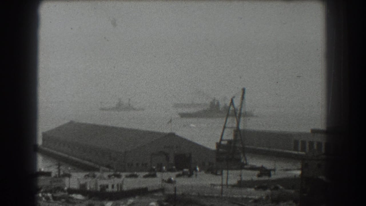 Hazy harbor view with distant ships, waterfront buildings, crane, and parked cars.