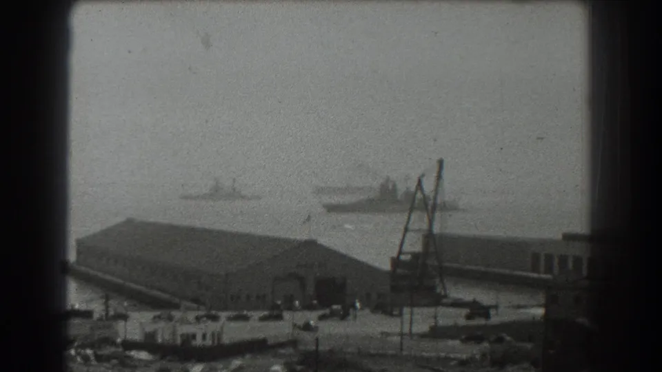 1939 San Francisco harbor view with distant ship silhouettes, dockside buildings, a crane, and parked cars seen through 8mm grain.