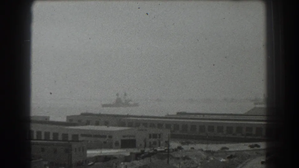 A distant vessel sits on hazy San Francisco Bay beyond low port buildings, a road, and scattered waterfront structures.