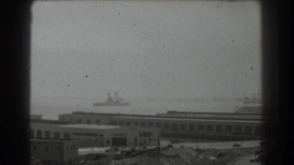 1939 San Francisco waterfront frame showing a long industrial building with a distant ship silhouette across the hazy bay.