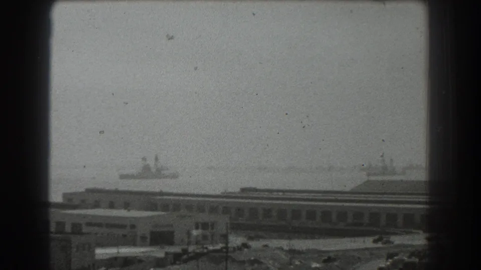 Hazy 1939 San Francisco harbor view with two distant ship silhouettes beyond low port buildings and open dockside ground.