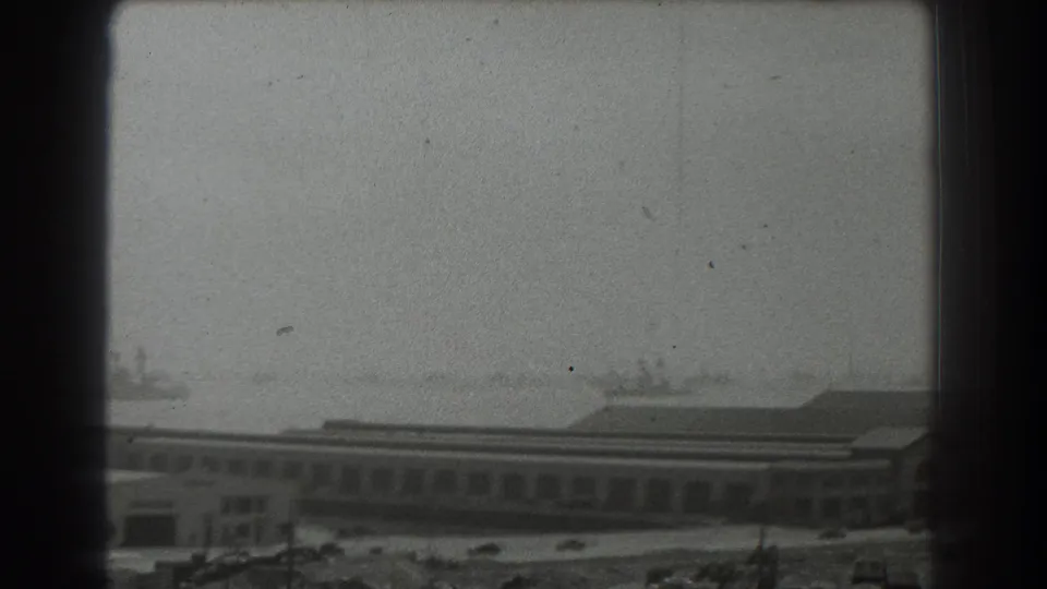 1939 home-movie frame of a hazy San Francisco harbor, with low waterfront structures and distant ships softened by grain.