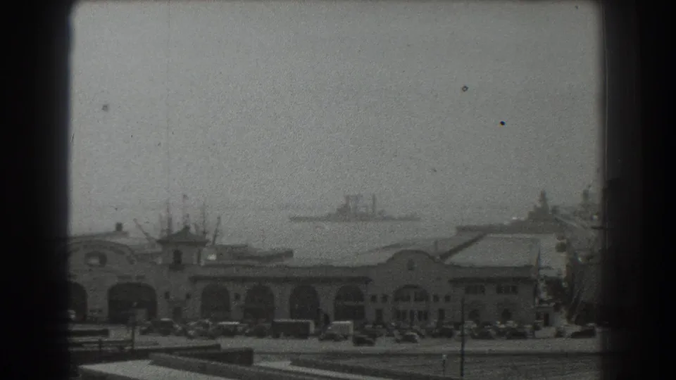 1939 San Francisco waterfront with arched terminal buildings, parked cars, open paved area, and a ship in the hazy distance.