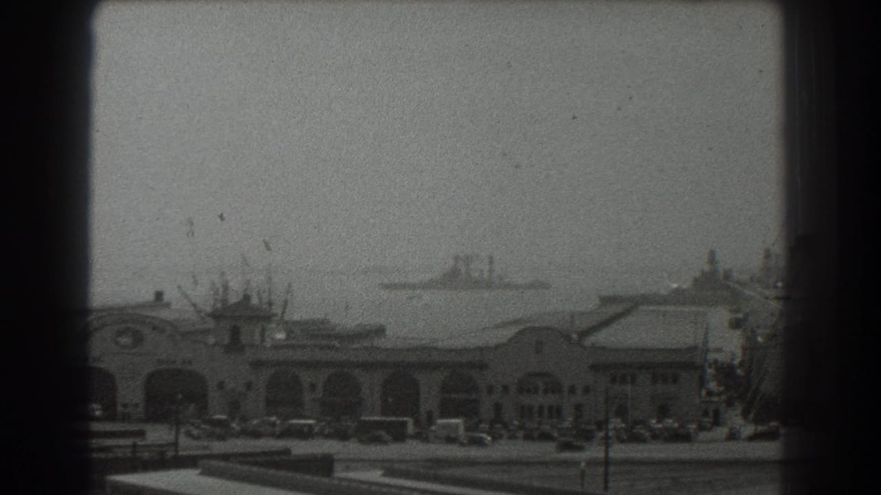 1939 San Francisco waterfront view with an arched pier building, parked cars, and distant ships on hazy bay water.