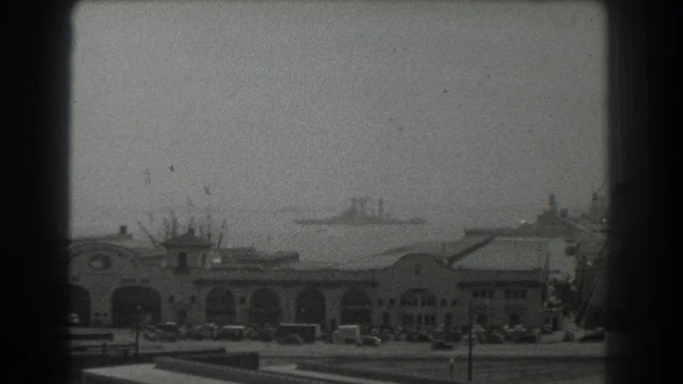 1939 San Francisco waterfront view with an arched pier building, parked cars, and distant ships on hazy bay water.