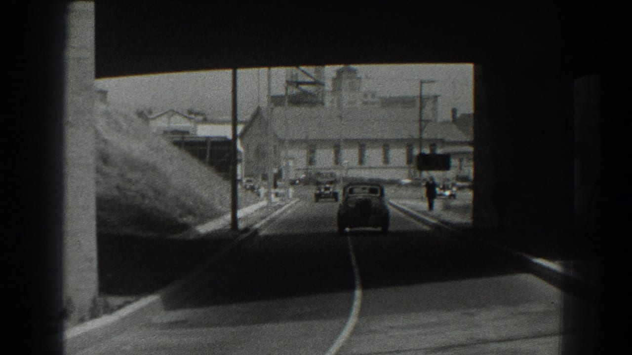 Vintage cars travel through a shadowed underpass toward sunlit industrial buildings.