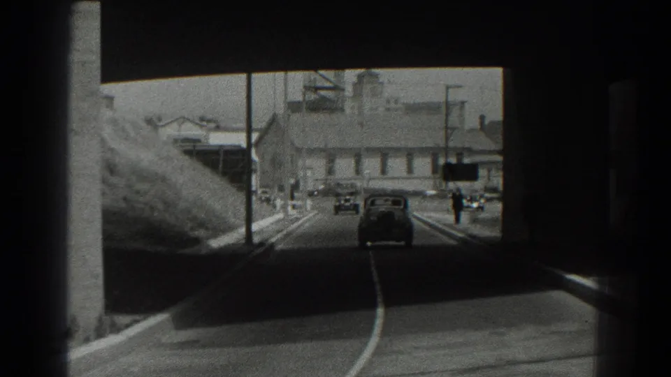 1939 San Francisco 8mm frame showing cars on a curving road beneath a dark bridge opening with hillside and industrial buildings ahead.