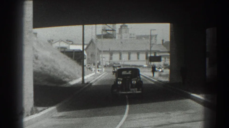 1939 San Francisco street view of a vintage car centered under an overpass, with bright industrial buildings and a small pedestrian beyond.