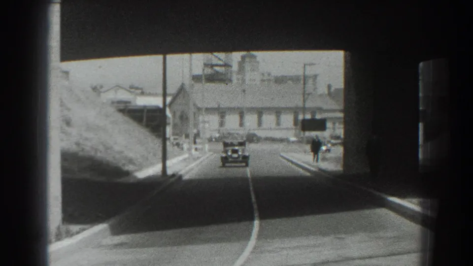1939 San Francisco 8mm frame showing a small car driving away from the shaded bridge area toward buildings, poles, hillside, and a distant pedestrian.