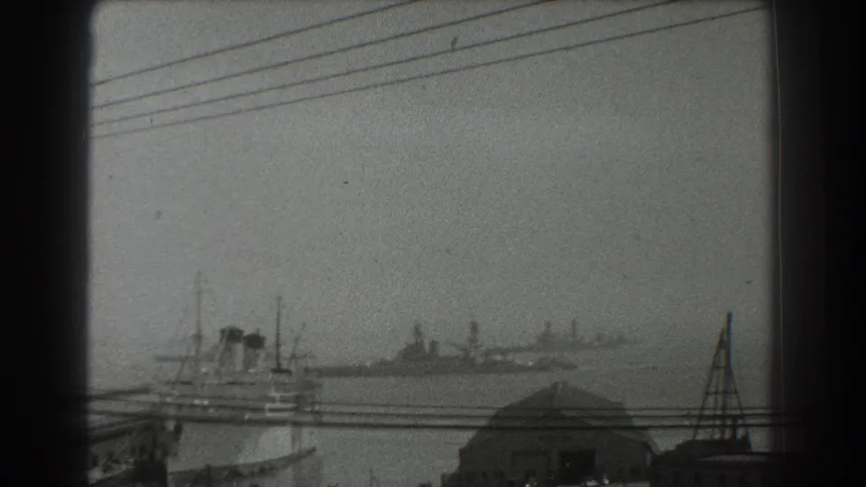 1939 San Francisco waterfront seen across hazy water, with large vessels, a dock roof, utility lines, and dark film-frame borders.