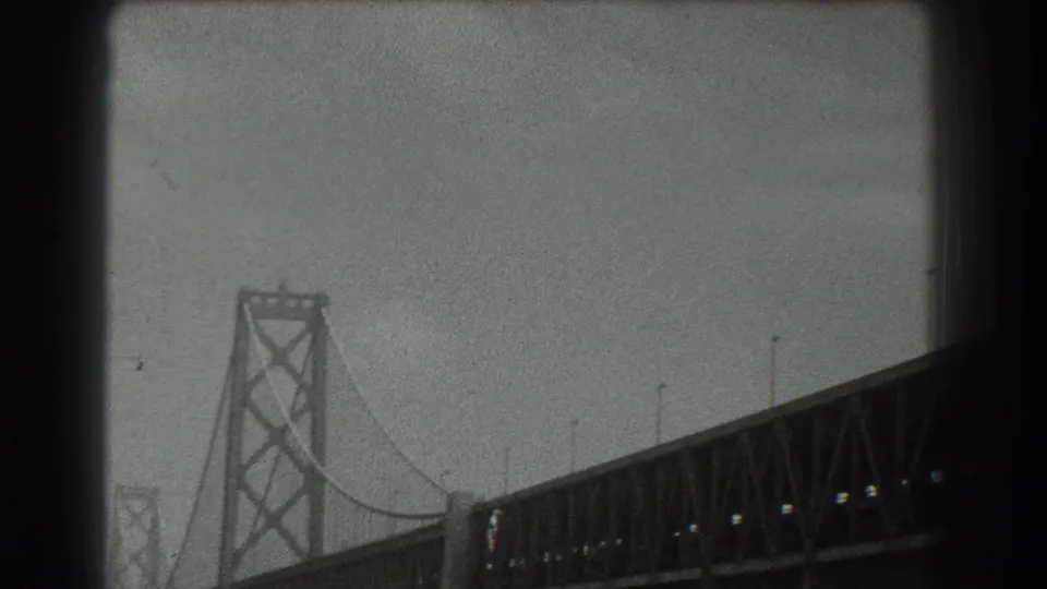 1939 8mm frame showing a Golden Gate Bridge tower, diagonal bracing, suspension cable, and bridge deck under overcast sky.