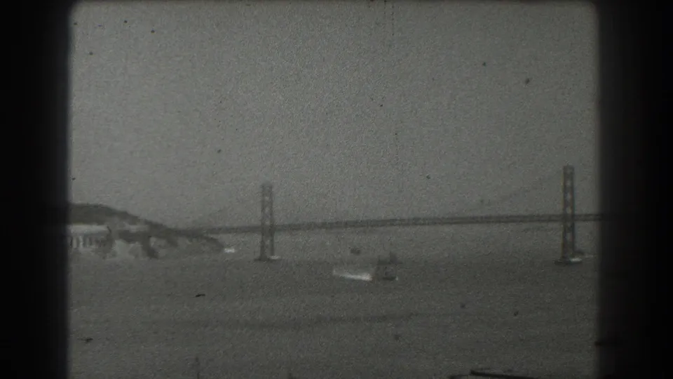 1939 San Francisco home-movie frame of the bridge crossing the bay, with water, shoreline, and small boat visible.