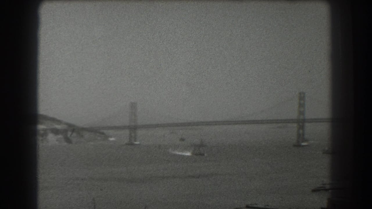 A 1939 San Francisco home-movie frame shows the Bay Bridge in the distance across hazy water, with small boats and wakes below the towers.