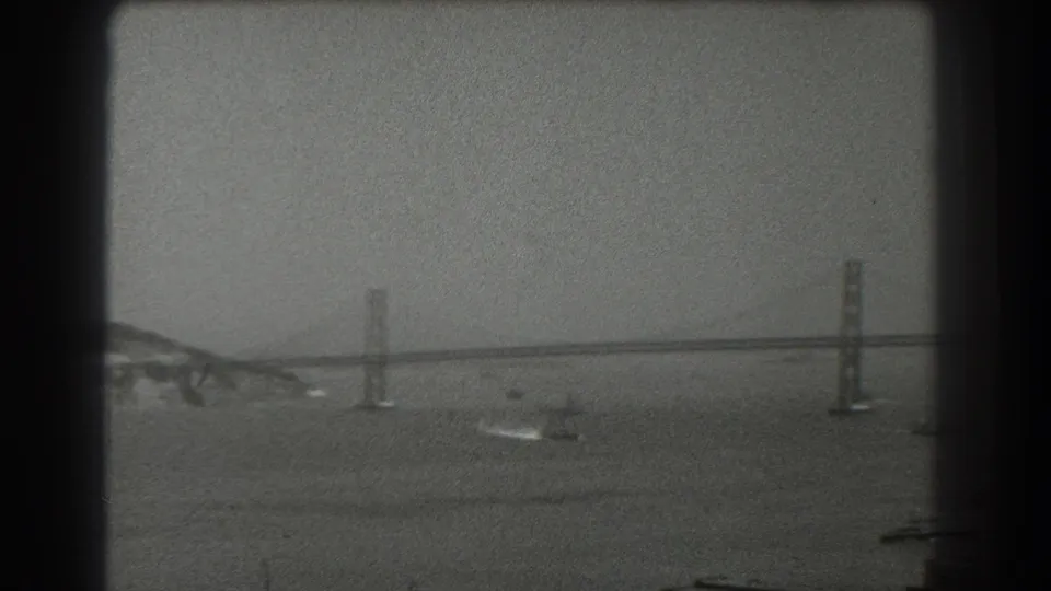 A 1939 San Francisco home-movie frame shows the Bay Bridge in the distance across hazy water, with small boats and wakes below the towers.