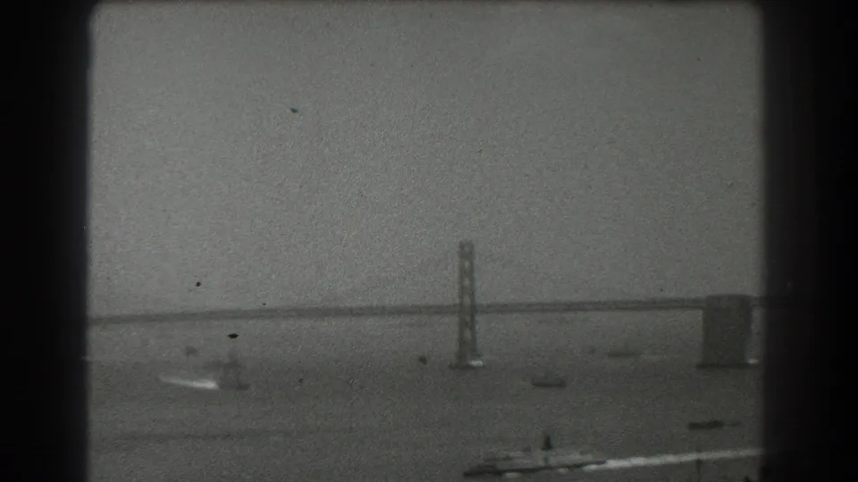 A soft 8mm frame from 1939 San Francisco shows a Bay Bridge tower and horizontal span across the water, with small boats scattered in the bay.