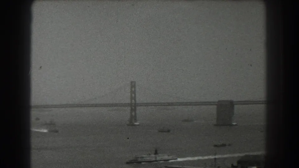A 1939 San Francisco bay scene shows a distant bridge tower and span above small boats, with film grain and dark frame edges visible.