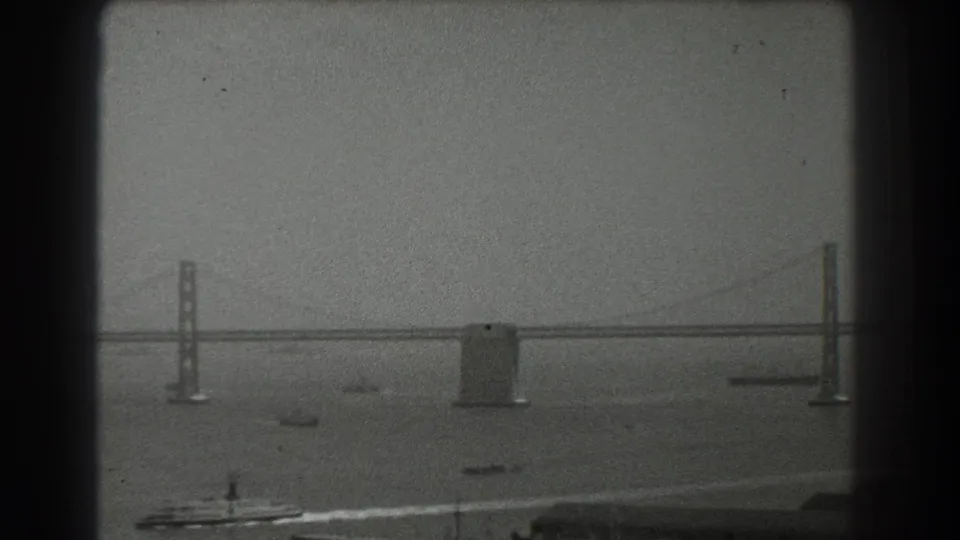 Black-and-white 8mm frame of the Bay Bridge crossing San Francisco Bay with boats moving below a hazy sky.