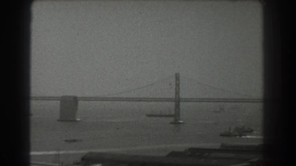 Late-1930s San Francisco Bay view with the Bay Bridge tower, long span, boats, haze, and dark shoreline along the lower frame.