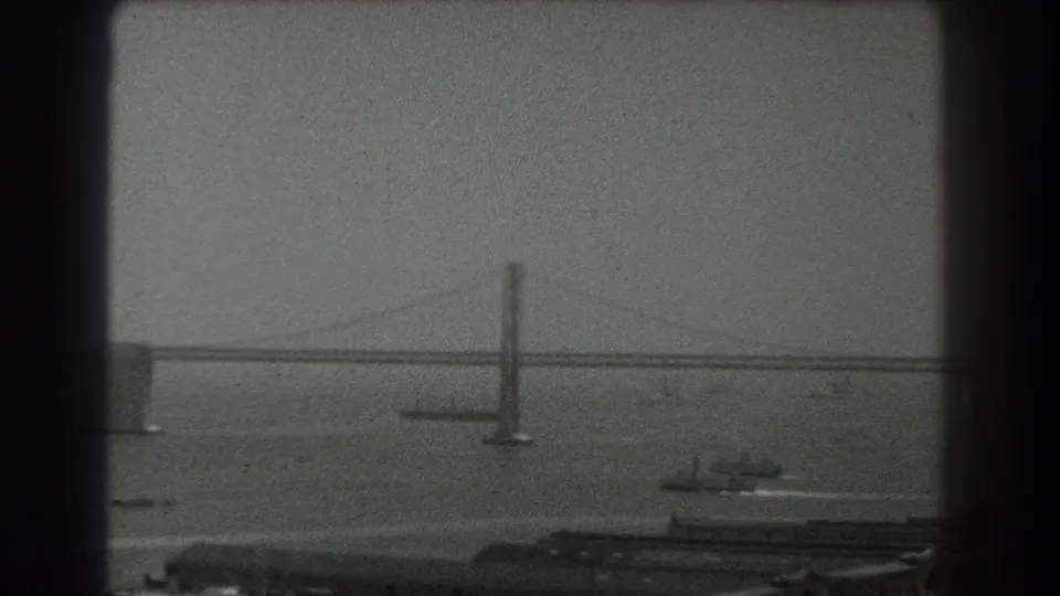 1939 San Francisco home-movie frame showing a hazy bay bridge span, tower, cables, small boats, and shoreline structures.