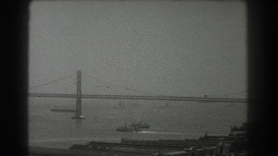 1939 San Francisco bay view with a suspension bridge tower, cable span, harbor boat wake, and muted sky.