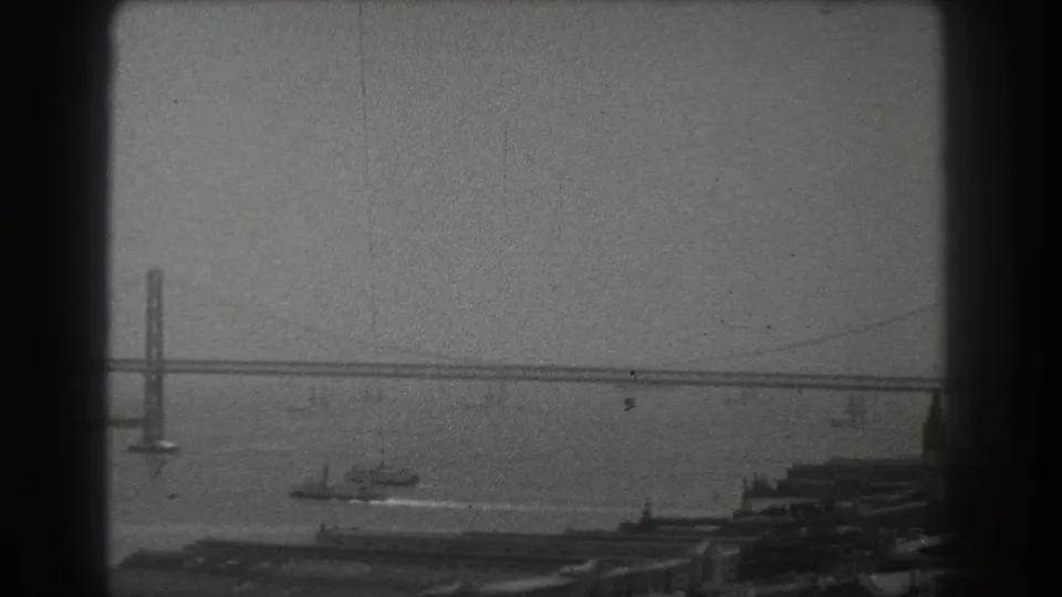 San Francisco 1939 home-movie frame with a bridge crossing the bay, a small vessel below, piers, and waterfront buildings.