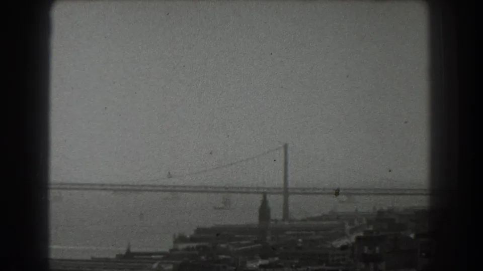 1939 San Francisco home-movie frame showing a suspension bridge across the bay above low waterfront structures.