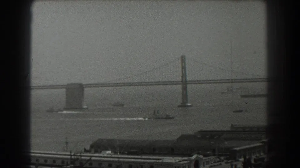 1939 San Francisco Bay frame showing bridge towers over the water, with small boats and industrial waterfront roofs below.