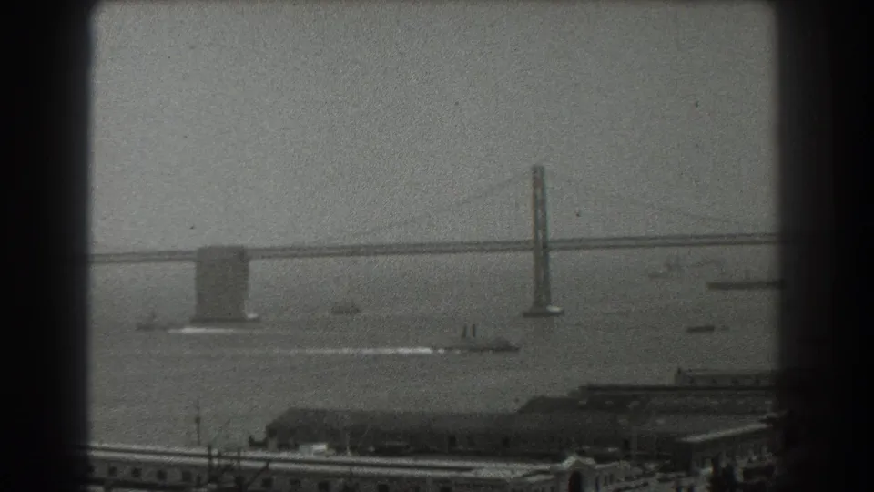 1939 San Francisco home-movie frame showing bridge towers, bay water, small vessels, and waterfront structures.