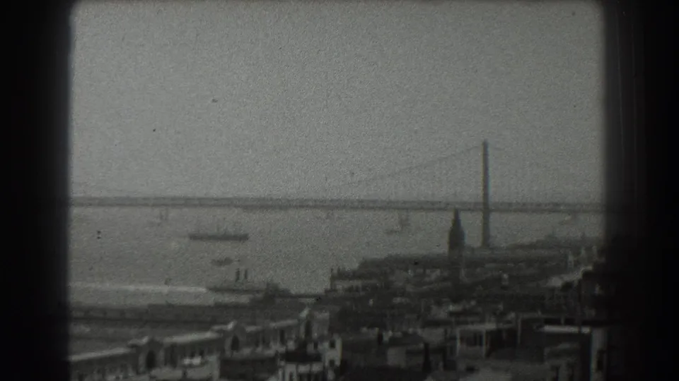 1939 8mm San Francisco Bay view with harbor rooftops, small vessels, a ferry wake, and the suspension bridge receding across the water.
