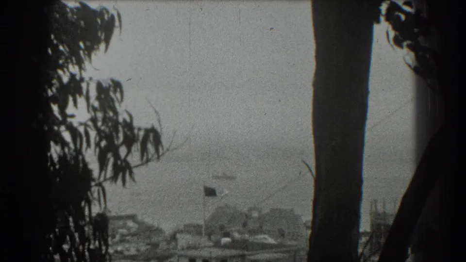 1939 San Francisco elevated view through dark trees, showing hazy water, a fluttering flag, low structures, and a distant ship.