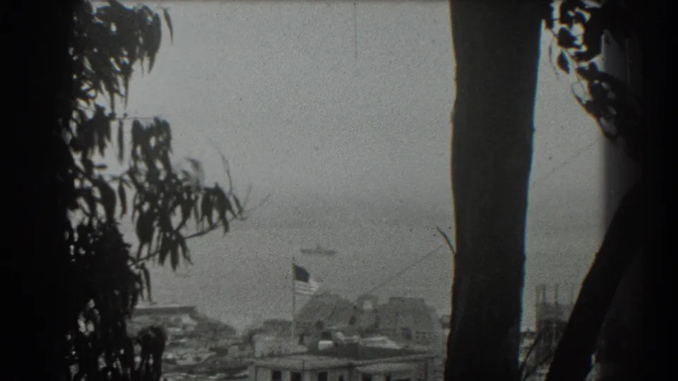 1939 San Francisco frame showing a tree-lined overlook toward hazy water, a distant ship, low waterfront buildings, and a small flag.