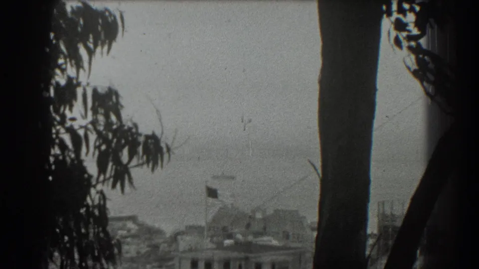 1939 San Francisco home-movie frame showing a bay and suspension bridge through dark foreground trees, with a rooftop flag at left.
