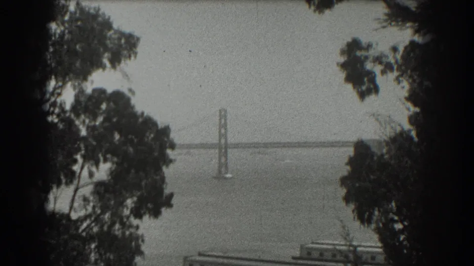 1939 San Francisco bay view with a suspension bridge tower and span framed by dark trees in grainy black-and-white 8mm footage.