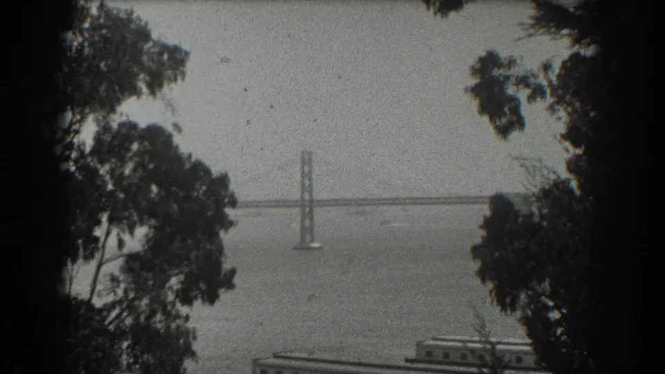 Elevated 1939 San Francisco home-movie view of a suspension bridge over the bay, framed by trees with a low waterfront structure below.