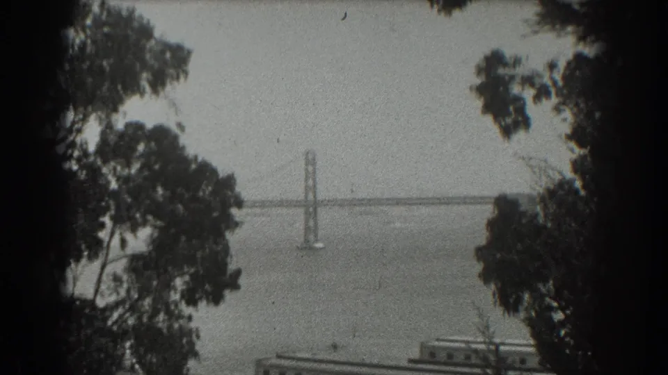 1939 San Francisco black-and-white 8mm frame of a suspension bridge across the bay, seen through trees from a high overlook.