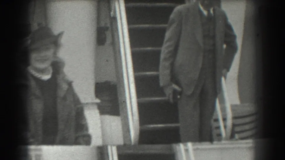A woman in a structured hat and coat moves through the foreground as a suited man stands near the staircase in 1939 San Francisco footage.
