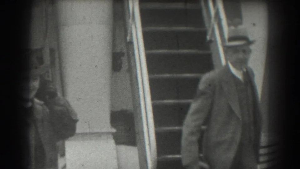The woman and the suited man appear near the lower part of the staircase, framed by rails and columns in black-and-white 8mm footage.