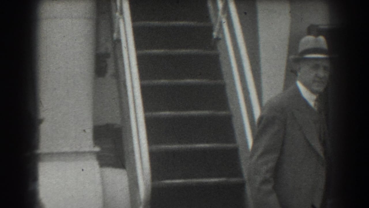 Man in a fedora and dark suit stands near stairs with bright metal railings.