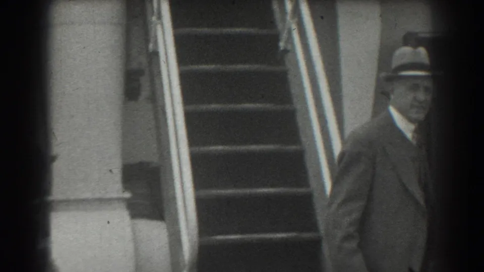 In 1939 San Francisco home-movie footage, a man in a fedora and dark suit is seen beside a staircase and wall.
