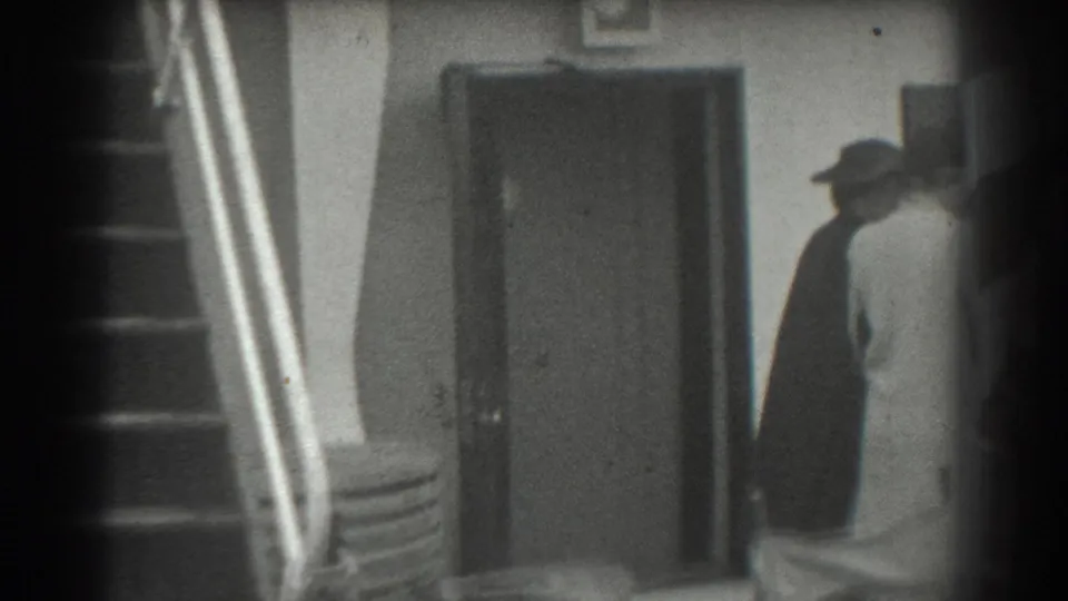 Two partially seen adult figures move near a closed doorway and metal-railed staircase in 1939 San Francisco home-movie footage.