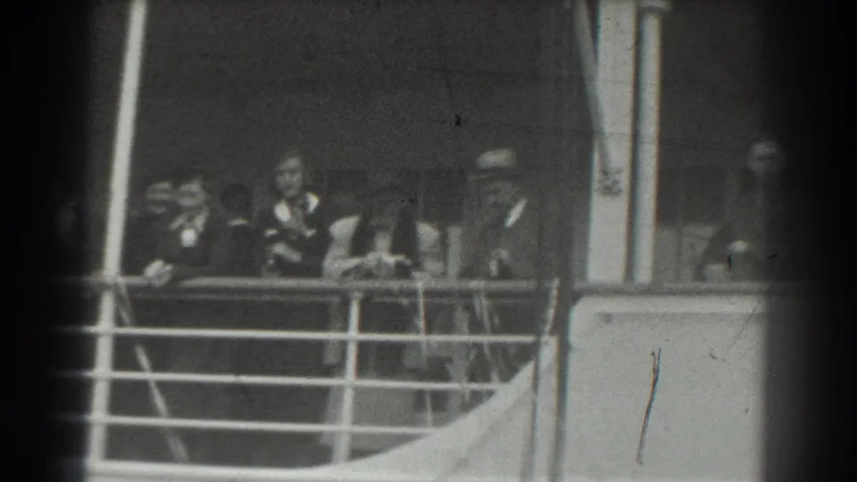 Black-and-white 8mm frame of ship passengers near the rail, with a deck wall and structural post visible.
