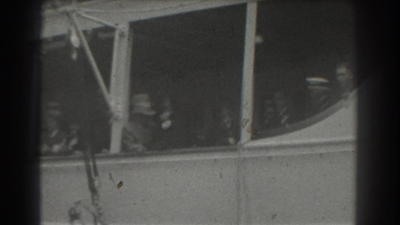 Passengers in hats visible through ship windows beside exterior ship structure.