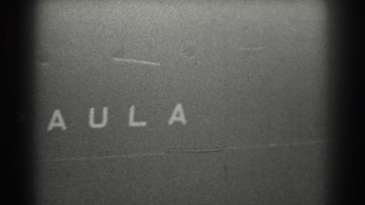 Black-and-white 8mm frame showing a partial title-card or leader fragment with the letters A U L A visible.