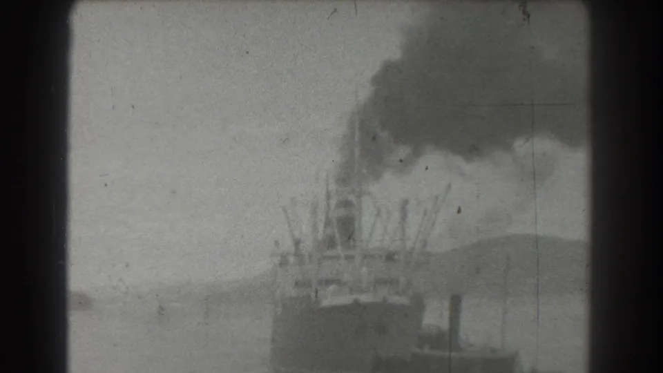 The 1939 harbor view holds on a steamship or industrial vessel releasing dark smoke, with a small tugboat close to the lower edge of the frame.