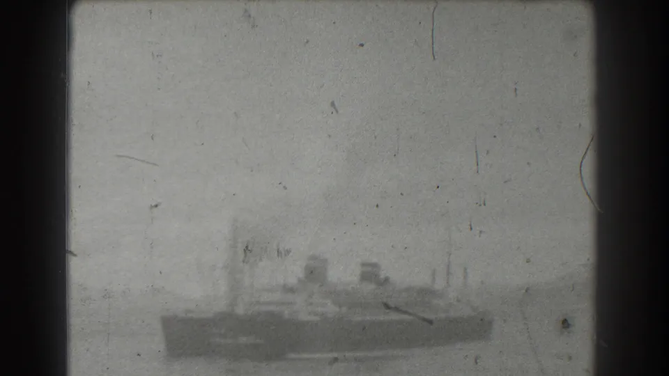 Black-and-white 8mm frame of a distant vessel moving across water near San Francisco, with a faint shoreline or hill form visible behind the stern.